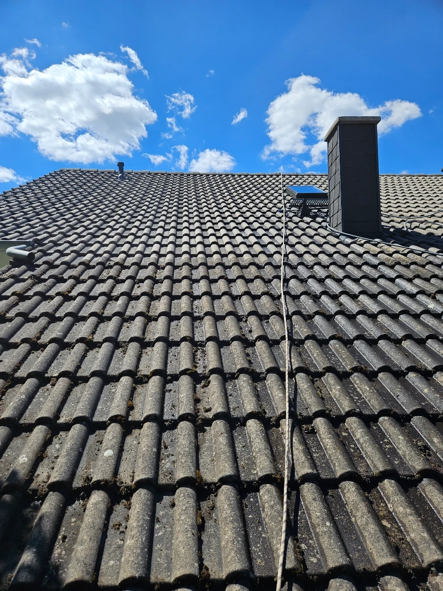 Nettoyage du toit : toiture en tuiles grises partiellement recouverte de mousse, avec une cheminée et un câble sous un ciel bleu lumineux