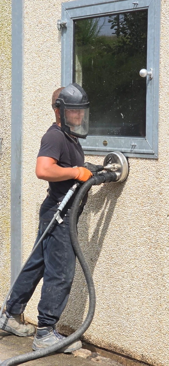 Nettoyage de la façade : opérateur masqué effectuant un ponçage aspirant sur une paroi extérieure rugueuse